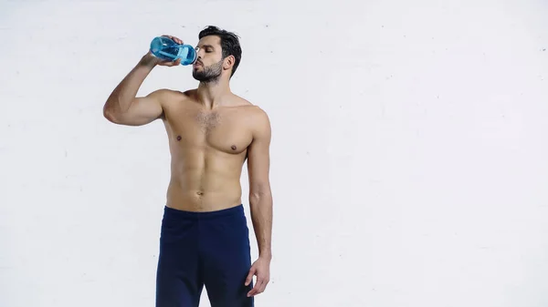 Deportista barbudo en pantalones cortos sosteniendo botella deportiva azul y agua potable cerca de la pared de ladrillo blanco - foto de stock