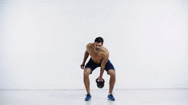 Full length of shirtless man exercising with heavy kettlebell near white brick wall — Stockfoto