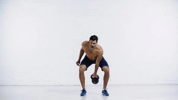 Comprimento total do homem sem camisa exercitando com kettlebell pesado perto da parede de tijolo branco — Fotografia de Stock