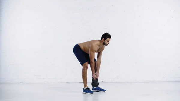 Homem barbudo e esportivo exercitando com kettlebell pesado perto da parede de tijolo branco — Fotografia de Stock