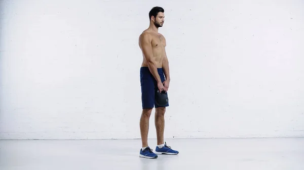 Full length of shirtless sportsman in shorts exercising with heavy kettlebell near white brick wall — Stock Photo