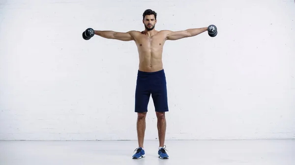 Full length of shirtless sportsman with outstretched hands holding heavy dumbbells near white brick wall — Fotografia de Stock