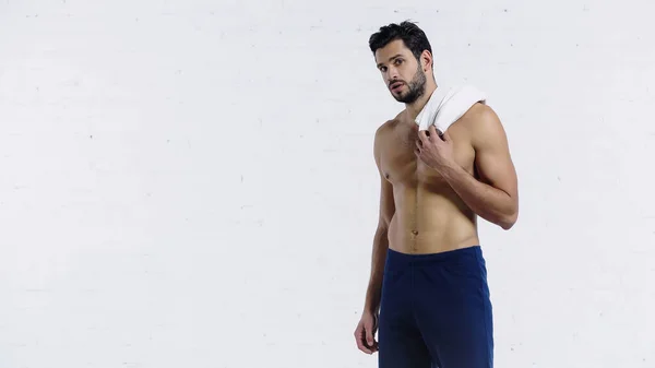 Deportista sin camisa sosteniendo la toalla y mirando a la cámara cerca de la pared de ladrillo blanco - foto de stock