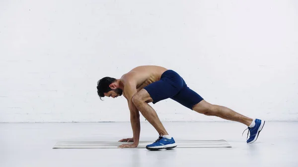 Vista lateral del deportista sin camisa haciendo ejercicio en la alfombra de fitness cerca de la pared de ladrillo blanco - foto de stock
