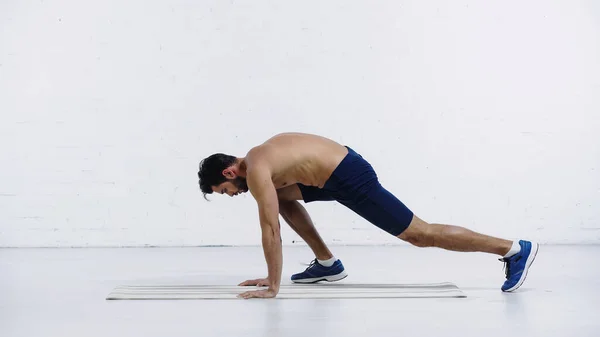 Side view of shirtless sportsman training on fitness mat near white brick wall — стоковое фото