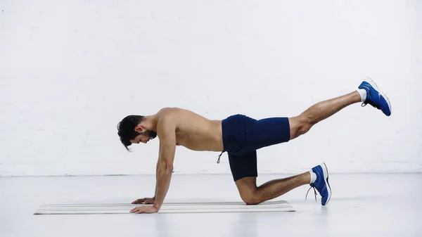 Vista lateral del deportista sin camisa haciendo tabla de la rodilla en la estera de fitness cerca de la pared de ladrillo blanco - foto de stock