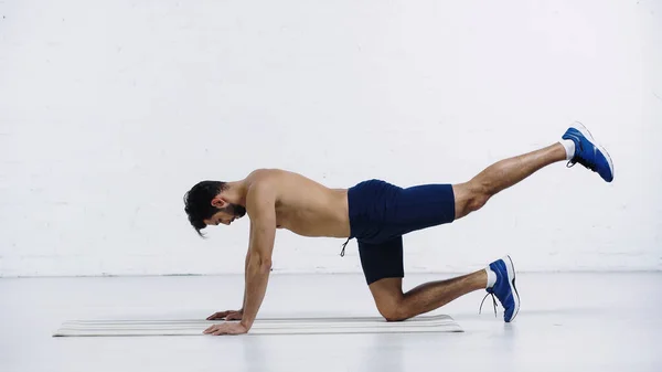 Side view of shirtless sportsman doing knee plank on fitness mat near white brick wall — стоковое фото