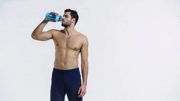 Deportista muscular en pantalones cortos azules beber agua de la botella de deportes cerca de la pared de ladrillo blanco - foto de stock