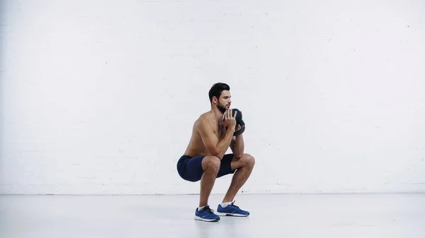 Full length of shirtless sportsman exercising with heavy kettlebell near white brick wall — Fotografia de Stock