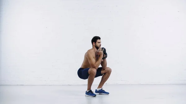 Full length of shirtless sportsman exercising with heavy kettlebell near white brick wall — стоковое фото