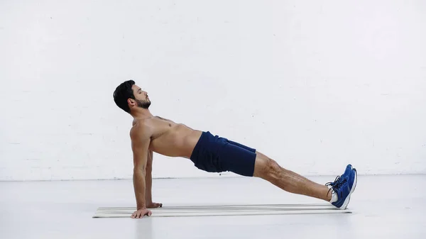 Vista lateral del deportista sin camisa en pantalones cortos y zapatillas de deporte de entrenamiento en la estera de fitness cerca de la pared de ladrillo blanco - foto de stock