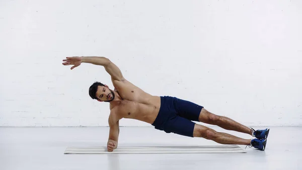 Shirtless sportsman doing side plank exercise on fitness mat near white brick wall — Stockfoto