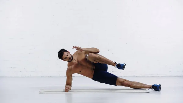 Desportista sem camisa fazendo exercício para pernas no tapete de fitness perto da parede de tijolo branco — Fotografia de Stock