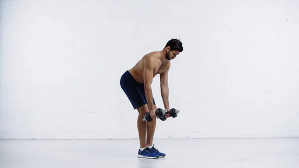 Desportista sem camisa no treinamento de shorts com halteres pesados perto da parede de tijolo branco — Fotografia de Stock