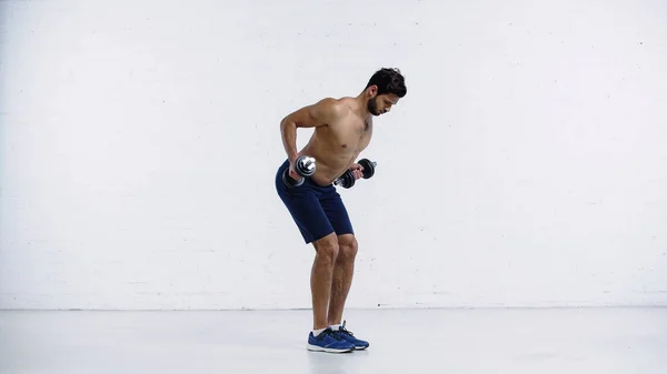 Полноразмерный спортсмен в кроссовках тренирующийся с тяжелыми гантелями возле белой кирпичной стены — стоковое фото