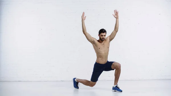 Toute la longueur du sportif musclé faisant des fentes avec les mains levées près du mur de briques blanches — Photo de stock