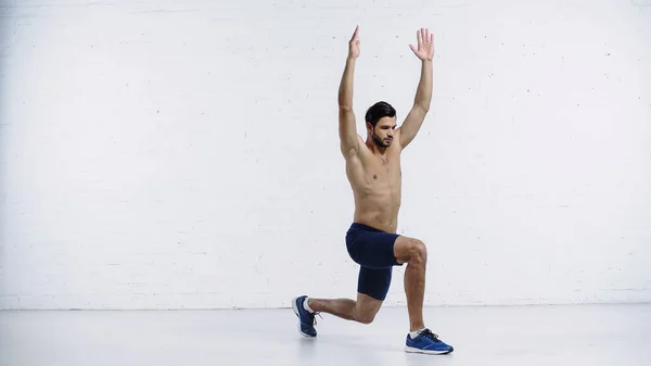 Full length of shirtless sportsman doing lunges with raised hands near white brick wall — Fotografia de Stock