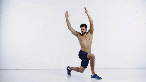 Longitud completa de barbudo joven deportista haciendo embestidas con las manos levantadas cerca de la pared de ladrillo blanco - foto de stock