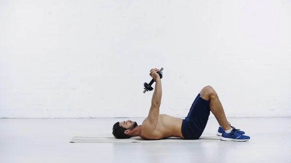 Seitenansicht des hemdslosen Sportlertrainings mit Kurzhantel im Liegen auf der Fitnessmatte in der Nähe der weißen Ziegelwand — Stockfoto