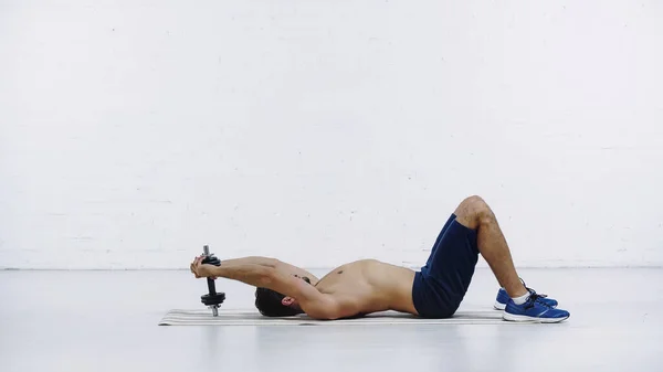 Visão lateral do desportista sem camisa no treinamento de shorts com halteres pesados enquanto deitado no tapete de fitness perto da parede de tijolo branco — Fotografia de Stock