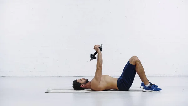 Visão lateral do desportista barbudo no treinamento de shorts com halteres pesados enquanto deitado no tapete de fitness perto da parede de tijolo branco — Fotografia de Stock
