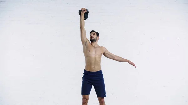 Muscular sportsman holding heavy kettlebell above head near white brick wall — Fotografia de Stock