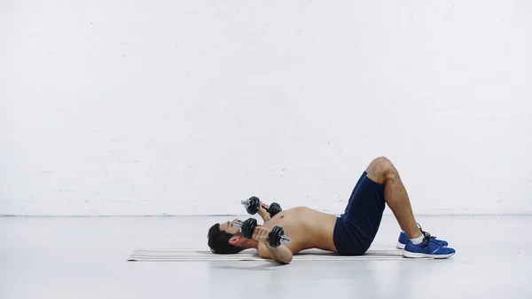 Vista lateral do homem sem camisa em shorts e tênis de treinamento com halteres enquanto deitado no tapete de fitness perto da parede de tijolo branco — Fotografia de Stock
