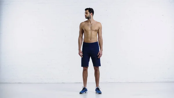 Full length of shirtless sportsman in shorts standing and doing neck exercise near white brick wall — Stock Photo