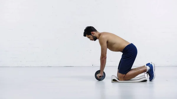 Side view of shirtless sportsman exercising with abdominal wheel near white brick wall — Stockfoto