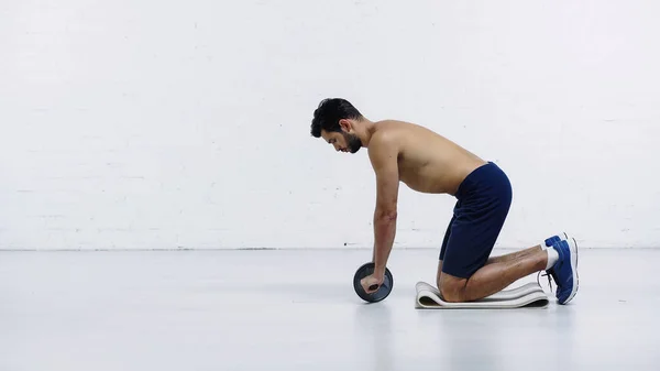 Vista lateral del deportista sin camisa haciendo ejercicio con rueda abdominal cerca de la pared de ladrillo blanco - foto de stock