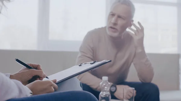 Psicólogo escribiendo en portapapeles cerca de pacientes borrosos y deprimidos hablando durante la consulta - foto de stock