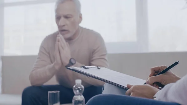 Psychologist writing on clipboard near blurred middle aged patient talking during consultation — Stock Photo