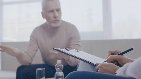 Psychologist writing on clipboard near blurred mature patient talking during consultation — Stock Photo