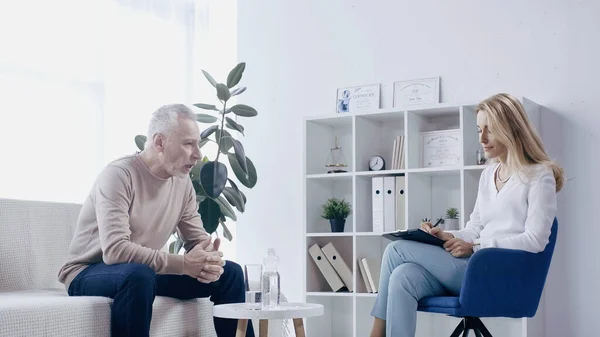 Mature man talking to psychologist with clipboard in consulting room — Fotografia de Stock