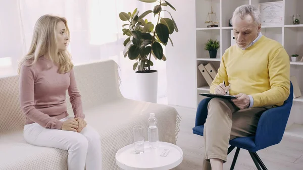 Mature psychologist writing on clipboard near blonde patient — Stock Photo