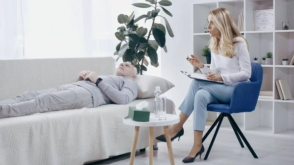 Middle aged sportsman lying on sofa and talking to blonde psychologist — Stock Photo