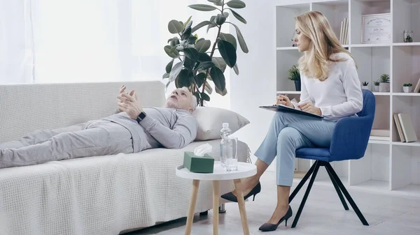 Mature man in sportswear lying on sofa and talking to blonde psychologist — Stock Photo