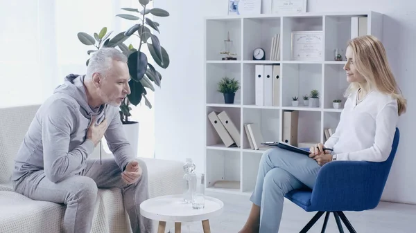 Homem maduro em sportswear sentado no sofá e falando com psicólogo loira — Fotografia de Stock
