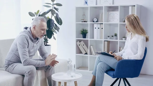 Homme mûr en vêtements de sport assis sur le canapé et avoir une conversation avec psychologue blonde — Photo de stock