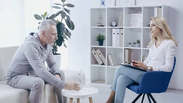 Mature man in sportswear taking glass of water near blonde psychologist — Foto stock