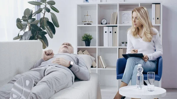 Hombre deprimido en ropa deportiva acostado en el sofá y hablando con el psicoterapeuta rubio con portapapeles - foto de stock