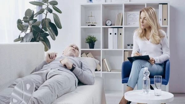 Hombre de mediana edad en ropa deportiva acostado en el sofá y hablando con el psicoterapeuta rubio con portapapeles - foto de stock