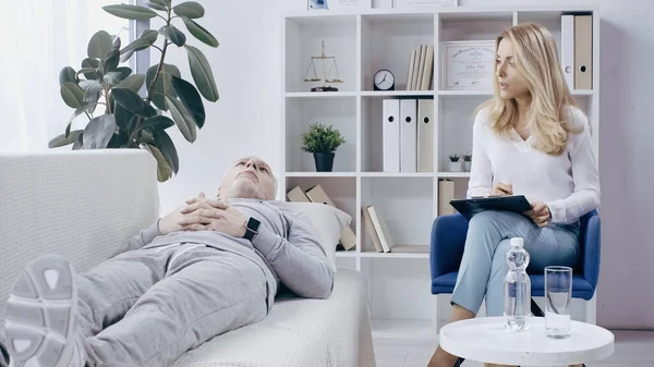 Blonde psychotherapist with clipboard talking to depressed man in sportswear lying on sofa in consulting room — Stockfoto