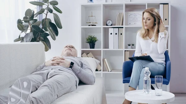 Blonde psychotherapist with clipboard talking to middle aged man in sportswear lying on sofa in consulting room — Stock Photo