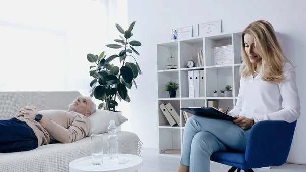 Blonde Psychotherapeutin schreibt auf Klemmbrett neben Mann mittleren Alters, der auf Couch im Beratungszimmer liegt — Stockfoto