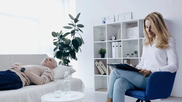 Psicoterapeuta rubia escribiendo en portapapeles cerca de hombre de mediana edad acostado en el sofá en la sala de consulta - foto de stock