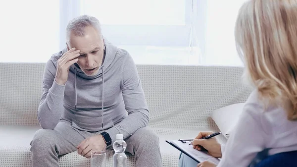 Чоловік середнього віку в спортивному одязі, що сидить на дивані біля розмитого психотерапевта в клініці — стокове фото
