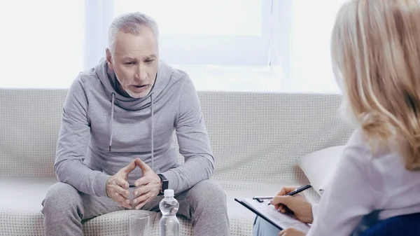 Sad middle aged man in sportswear sitting on couch near blurred psychotherapist in clinic — Stock Photo