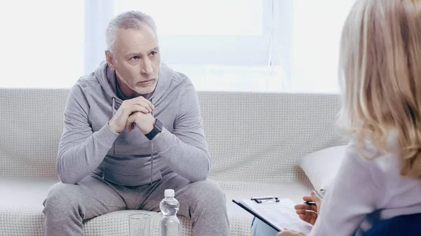 Middle aged man in sportswear sitting on couch and looking at blurred psychotherapist in clinic — Fotografia de Stock