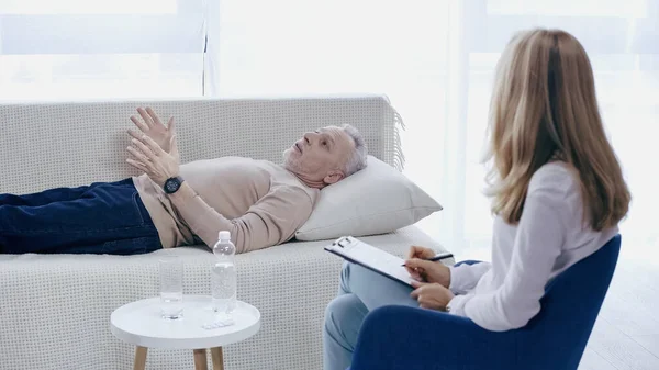 Psicoterapeuta sosteniendo pluma cerca del portapapeles y escuchando hombre de mediana edad acostado en el sofá en la sala de consulta - foto de stock
